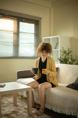 Rahatlamış bir kadın sabah kahvesinin tadını çıkarıyor ve telefonuna hafifçe aydınlatılmış bir evde bakıyor..