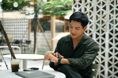 Asyalı adam dışarıdaki kafe masasında otururken etrafı kolaçan ediyor. Yanında buzlu kahve ve dizüstü bilgisayarla..