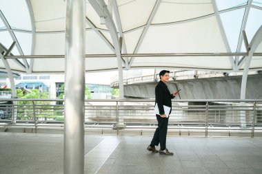 Asyalı işadamı, elinde akıllı telefon ve dizüstü bilgisayarla modern geçiş yolunda yürüyor..