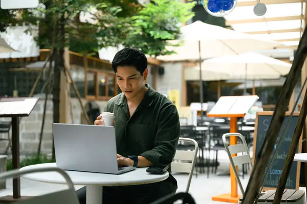 Gülümseyen adam kahve içiyor ve modern bir açık hava kafede dizüstü bilgisayar kullanıyor..