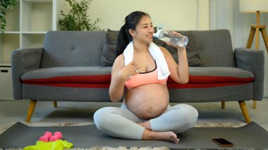 Mutlu hamile kadın egzersizden sonra dinleniyor, yoga minderinde oturuyor ve su içiyor..
