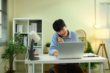 Masada dizüstü bilgisayarla çalışırken telefonla konuşan mutlu genç girişimci..