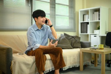 Evdeki kanepede oturan genç bir adam, rahat bir oturma odasında telefon görüşmesi yapıyor..