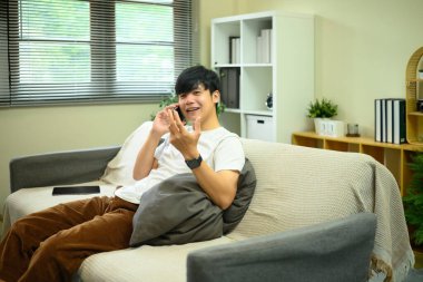 Evdeki kanepede oturan genç bir adam, rahat bir oturma odasında telefon görüşmesi yapıyor..