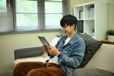 Oturma odasında dijital tablet kullanan kot ceket giyen mutlu Asyalı genç adam..