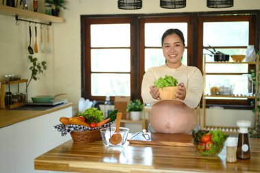 Asyalı hamile kadın parlak bir mutfakta duruyor, bir kase salata sunuyor..
