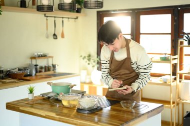 Asyalı bir adam yemek yaparken yemek tarifini kontrol ediyor ya da not alıyor..
