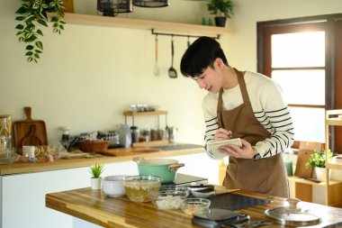 Asyalı bir adam yemek yaparken yemek tarifini kontrol ediyor ya da not alıyor..