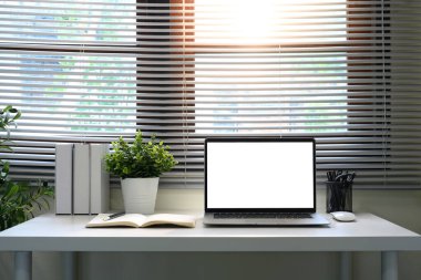 Boş bir dizüstü bilgisayar, defter ve pencere kenarındaki bitkilerle minimalist çalışma alanı.