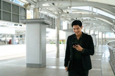 Genç bir erkek yönetici modern bir yürüyüş yolunda yürüyor ve akıllı telefonunu kontrol ediyor..