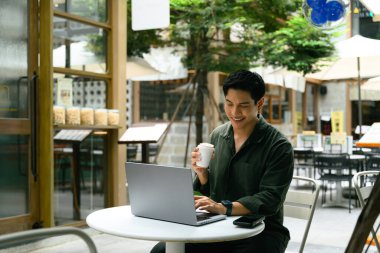 Açık bir kafede dizüstü bilgisayarda çalışan ve bir fincan kahvenin keyfini çıkaran gülümseyen adam..