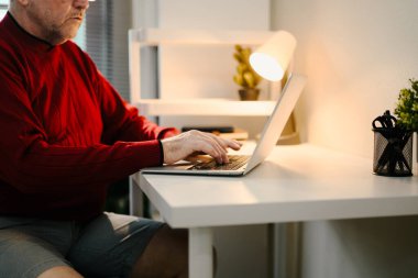 Masa lambasının altındaki minimalist masada dizüstü bilgisayar kullanan yaşlı adamı kapat..