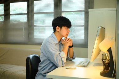 Genç adam evde çalışıyor, masada oturuyor ve masaüstü bilgisayar ekranına konsantre oluyor..