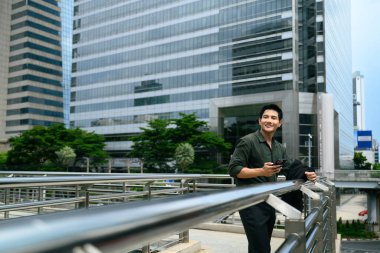 Güler yüzlü iş adamı elinde akıllı telefonuyla şehir merkezinde parmaklıklara yaslanıyor..