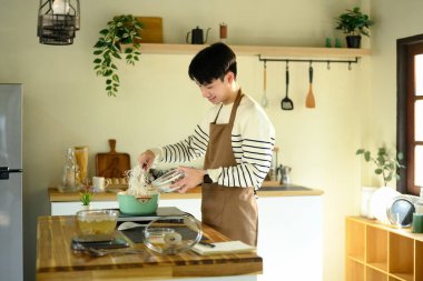 Önlüklü gülümseyen adam güneşli bir mutfakta tencereye erişte ekliyor..