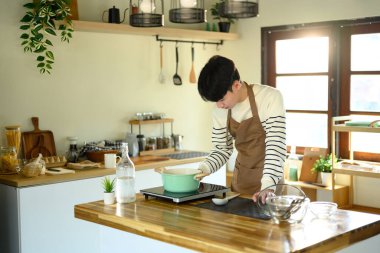 Güneşli, modern bir mutfakta yemek hazırlayan önlüklü, gülümseyen adam..