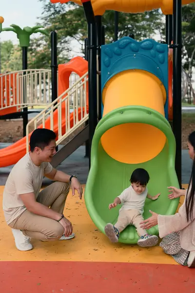 Mutlu ebeveynler, çocuklarının renkli bir kaydırağın kayışını izleyip alkışlıyorlar..