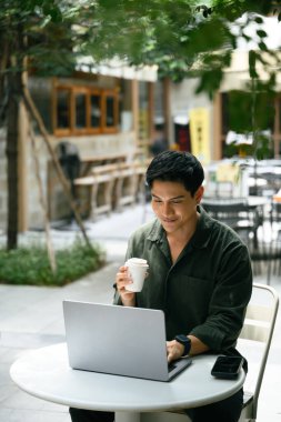 Yetişkin bir adam, modern bir açık hava kafede laptopuyla çalışırken kahveden zevk alıyor..