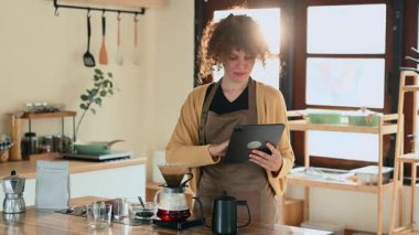 Genç bir kadın kahveyle demleniyor ve şık bir mutfakta dijital tablet kullanıyor..