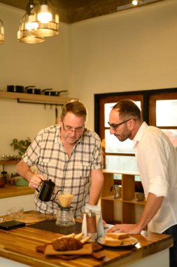 Kıdemli adam ve yetişkin oğlu ahşap mutfak tezgahında el yapımı kahve hazırlıyorlar. Babalar Günü konsepti.