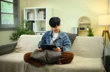 Kot ceketli bir adam dijital tablet kullanıyor. Oturma odasındaki koltukta uzanıyor..