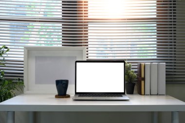 Boş bir ekran dizüstü bilgisayarı, kahve kupası, kitaplar ve pencere kenarındaki bitkilerle merkez ofis masası...