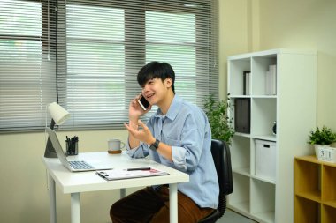 Masada oturan gülümseyen adam dizüstü bilgisayarı üzerinde çalışırken akıllı bir telefonla konuşuyor..