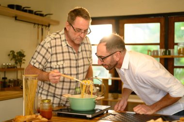 Mutlu kıdemli adam ve yetişkin oğlu parlak bir mutfakta makarna yemeği hazırlıyor. Babalar Günü konsepti.