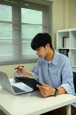 Genç işadamı, açık bir dizüstü bilgisayarın yanındaki kağıt üzerindeki veri grafiklerini ve grafikleri inceliyor..