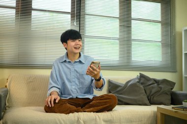 Evdeki kanepede oturan gülümseyen genç adam, elinde akıllı bir telefonla sohbet ediyor..