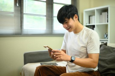 Rahat bir oturma odasında otururken akıllı telefon kullanan gülümseyen adam..