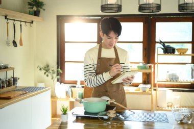 Önlüklü genç adam parlak bir mutfakta yemek tarifi ya da yemek planı yazıyor..