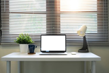 Bilgisayarı, kahve fincanı, iç mekan bitkileri ve pencerenin altında lamba olan parlak bir ofis..