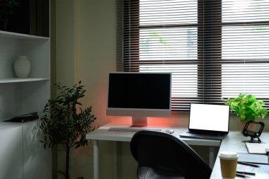 Masaüstü monitörü, dizüstü bilgisayarı ve pencere perdelerinin yanındaki yeşillik ile modern ofis kurulumu.