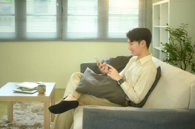 Kanepede oturan genç adam, parlak bir oturma odasında akıllı telefonunu kullanıyor..