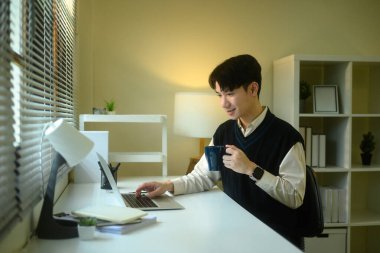 Gülümseyen Asyalı adam bir ofiste dizüstü bilgisayar kullanırken sıcak bir içkinin tadını çıkarıyor..