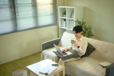 Evden uzakta bir kanepede dizüstü bilgisayarla çalışan ve elinde kahve fincanı tutan genç bir adam..