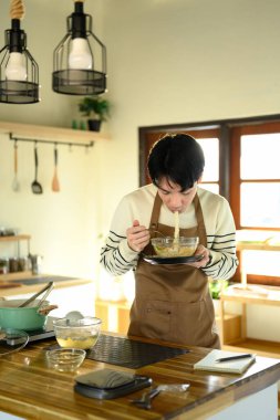 Önlüklü Asyalı bir adam cam bir kaseden erişte yiyor, bir ev mutfağında duruyor..