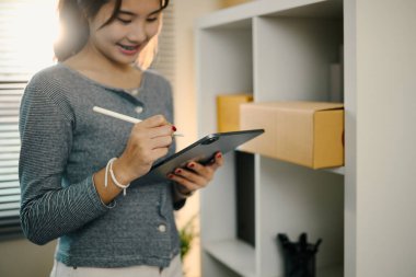 Genç girişimci, kargo kutularıyla rafların yanında online iş siparişlerini yönetmek için dijital tablet kullanıyor.