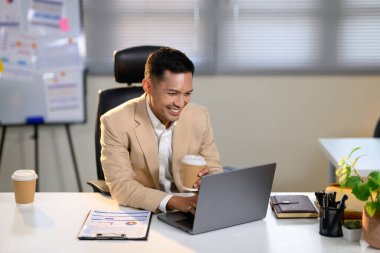 Gülümseyen işadamı, modern ofiste dizüstü bilgisayarla çalışırken elinde kahve fincanı tutuyor..
