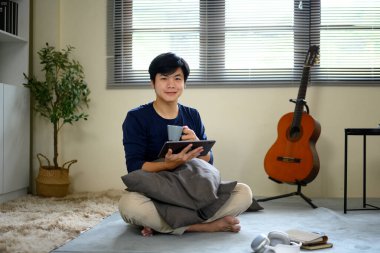 Evde elinde dijital tablet ve kahve fincanı tutarak rahatlayan genç adam yerde oturuyor..