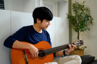 Genç Asyalı adam evde dinleniyor ve akustik gitar çalıyor..