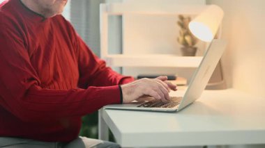 Kırmızı kazaklı son sınıf öğrencisi doğal ve lamba ışığında sakin bir odada çalışıyor..