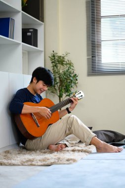 Genç adam rahat bir oturma odasında dinlenirken akustik gitar çalıyor..