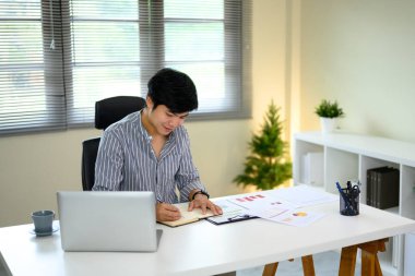 İşadamı masa başında dizüstü bilgisayar ve belgelerle çalışırken notlar yazıyor..