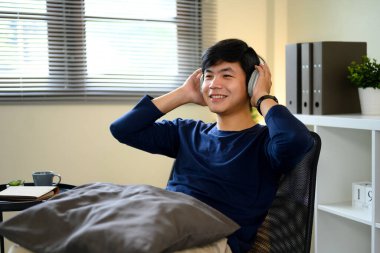 Modern bir ofiste oturup, kablosuz kulaklıkla müzik dinleyen mutlu genç bir adam..