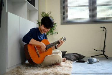 Rahat giysiler içinde, salonda yerde gitar çalan mutlu Asyalı..