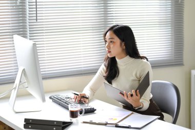 Masasında elinde bir dosya ve masaüstü bilgisayarıyla çalışan kendinden emin genç bir kadın..