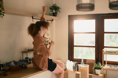 Genç bir kadın elinde bir fincan kahve tutuyor ve güneşli bir mutfakta neşeyle esniyor..