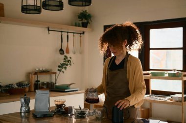 Önlüklü genç bir kadın güneşli bir mutfakta kahve hazırlıyor..